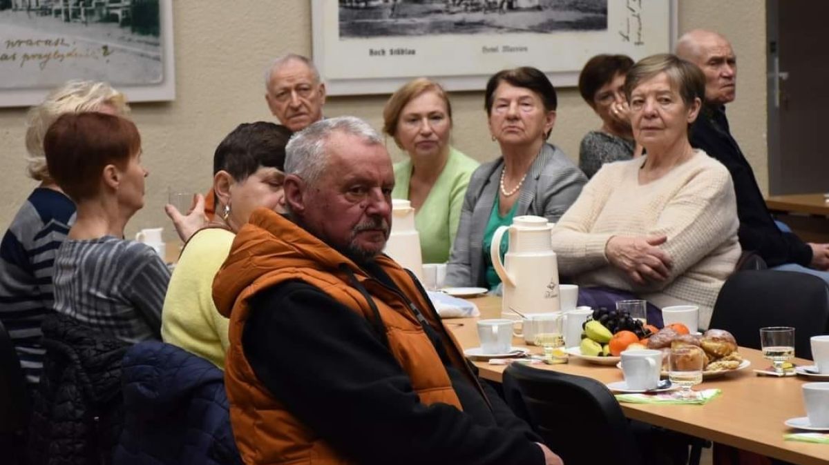 zdjęcie przedstawia ludzi siedzących przy stole podczas zebrania seniorów na sali